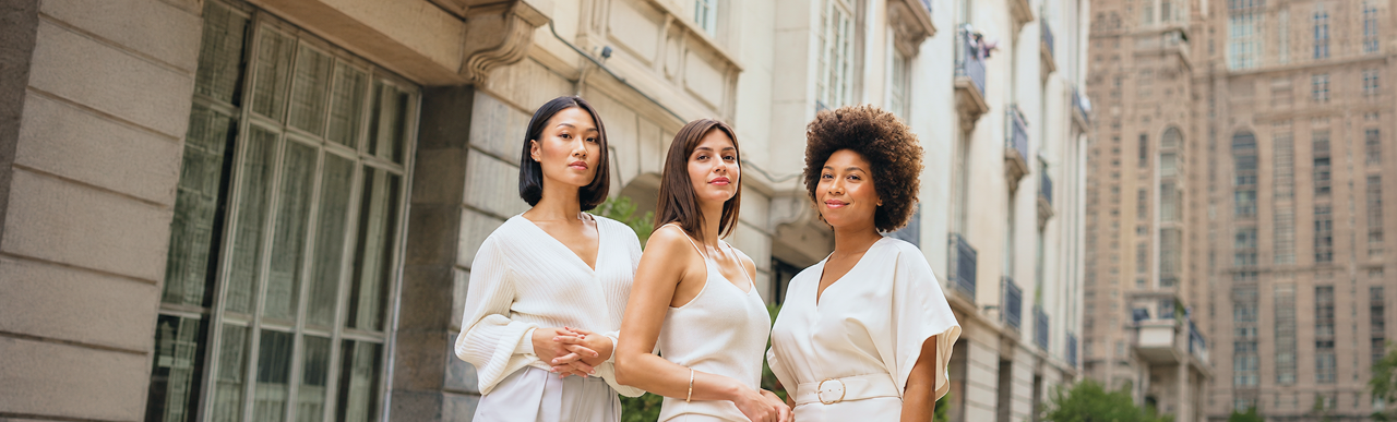 Três mulheres de etnias diferentes, lado a lado, vestindo roupas em tons de branco, olhando diretamente para a câmera com expressão séria. Ao fundo, fachadas de prédios clássicos em uma rua urbana.
