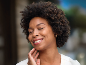 Mulher negra de cabelo cacheado, sorrindo com os olhos fechados, com a mão tocando o queixo. Ela usa brincos pequenos e camisa branca. Ao fundo, ambiente externo desfocado.