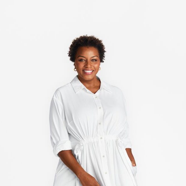 Mulher negra de cabelo curto cacheado, sorrindo, usando brincos e vestindo camisa-vestido branca com cinto na cintura. Fundo neutro claro.