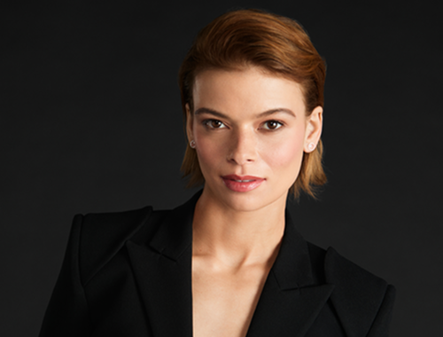 Mulher de cabelo curto ruivo-acobreado, penteado para trás, com expressão séria e leve sorriso, vestindo blazer preto. Fundo escuro e neutro.