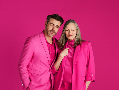 Homem e mulher lado a lado, ambos vestidos em conjuntos rosa-pink, olhando para a câmera com expressão confiante. Fundo em rosa-magenta vibrante.