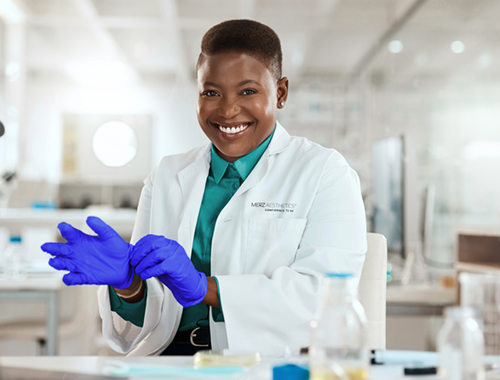 Mulher negra de cabelo curto, sorrindo, vestindo jaleco branco da Merz Aesthetics e calçando luvas azuis. Ao fundo, laboratório com frascos e equipamentos.