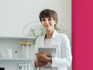Mulher de cabelo curto castanho, vestindo jaleco branco, segurando um tablet e sorrindo. Ao fundo, prateleiras com frascos e uma planta, com faixa vertical rosa-escuro ao lado.