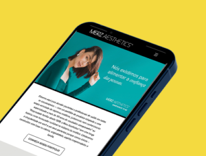 Smartphone exibindo página do site Merz Aesthetics em fundo verde-azulado, com foto de mulher sorrindo tocando o rosto, texto ao lado e um botão "Conheça nosso portfólio" abaixo. Fundo da imagem em amarelo.