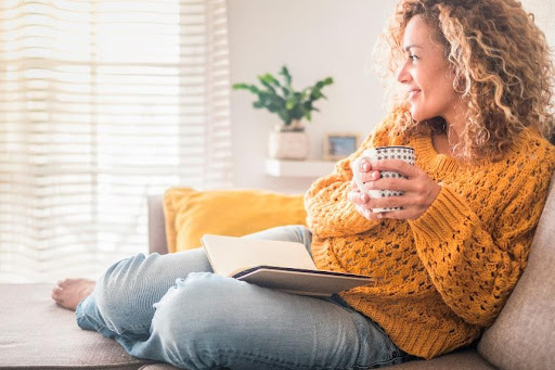 Mulher de cabelo cacheado ruivo, sentada no sofá com um livro aberto no colo, segurando uma xícara e olhando pela janela. Ela veste suéter amarelo mostarda e calça jeans. Ao fundo, planta e cortinas claras.