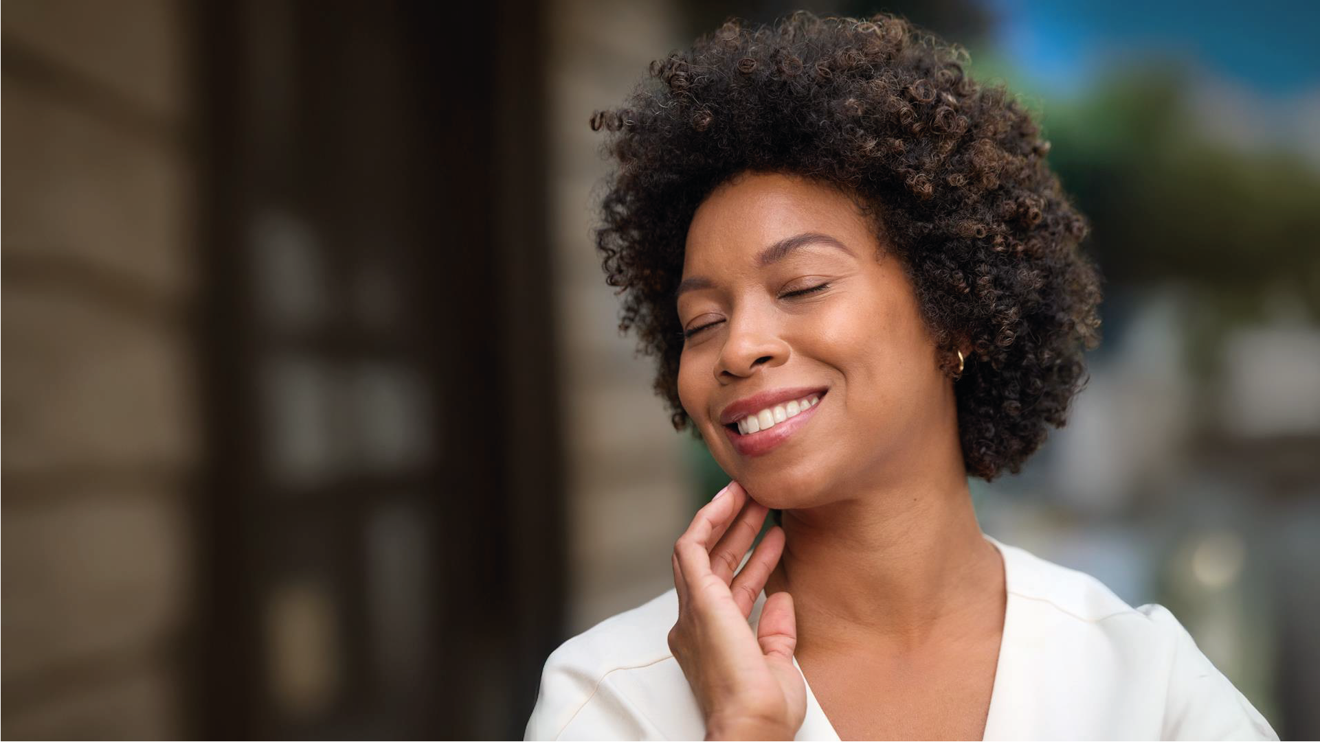 Mulher negra de cabelo cacheado, sorrindo com os olhos fechados, com a mão tocando o pescoço, vestindo blusa branca. Ao fundo, ambiente externo desfocado com prédio.
