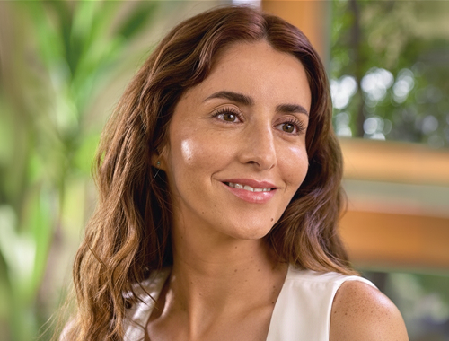 Mulher de cabelo longo e ondulado castanho, sorrindo levemente e olhando para o lado, vestindo blusa branca. Ao fundo, ambiente com plantas e estrutura de madeira desfocados.