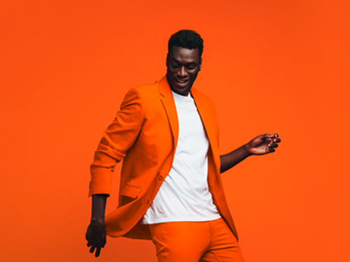 Homem negro sorrindo, em pose animada de dança, vestindo terno e calça em tom laranja sobre camiseta branca. Fundo laranja vibrante.